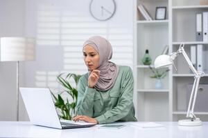 uma dedicada profissional vestindo uma hijab é profundamente focado Como ela trabalho em dela computador portátil às uma brilhante escritório escrivaninha. foto