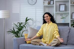 jovem lindo mulher meditando às casa sentado em sofá dentro vivo quarto dentro lótus pose, latim americano Veja Câmera sorridente, sonhando e visualizando futuro realização resultados e planos. foto