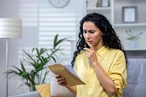 chateado e triste mulher às casa recebendo mau notícia notificação carta, hispânico mulher sentado em sofá dentro vivo quarto com enviar envelope nervosamente leitura. foto