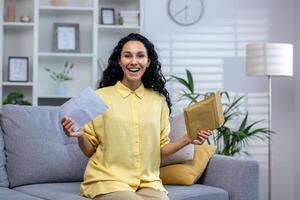 feliz cabelo encaracolado hispânico mulher sentado às casa em sofá dentro vivo sala, segurando aberto envelope. fofa morena é feliz com a notícias, ganhando loteria, feliz notícias, sorridente e olhando às a Câmera. foto