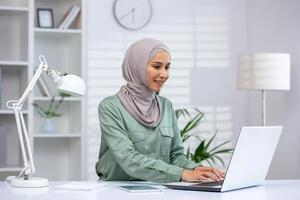 uma muçulmano mulher dentro uma hijab trabalho diligentemente em dela computador portátil às uma bem organizado escritório mesa, cercado de luz e moderno decoração. foto
