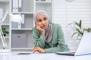 uma jovem meio Oriental mulher vestindo uma hijab senta às dela escrivaninha dentro a escritório, aparecendo pensativo e pensativo com uma computador portátil e caderno dentro frente do dela. foto