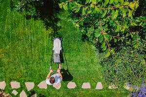 caucasiano homem empurrando gramado cortador de grama para corte verde Relva dentro jardim com luz solar às verão temporada. aéreo visualizar. tarefas domésticas e estilo de vida conceito. foto