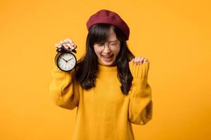 dentro dela chique escritório, uma jovem ásia mulher, adornado dentro uma amarelo suéter e vermelho boina, sorrisos calorosamente de a alarme relógio, exibindo dela pontualidade. foto