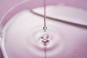 sérum ou Cosmético óleo fluxos para dentro uma transparente tigela em uma roxa fundo. foto