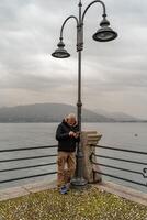 atraente meia idade homem usando Móvel telefone em pé perto uma rua luminária em a beira do lago dentro Stresa foto