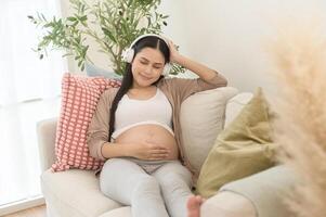 feliz grávida mulher com fones de ouvido ouvindo para Mozart música e deitado em sofá, gravidez conceito foto