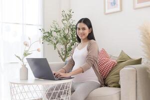 grávida mulher trabalhando em computador portátil e inteligente telefone dentro a vivo quarto às casa foto
