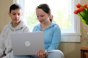 menina e Garoto adolescentes fazendo dever de casa ou assistindo filmes em computador portátil sentado em pequeno sofá contra janela Garoto mordeu lábio olhando atentamente menina mostra sorridente às monitor tela real pessoas vida estude foto