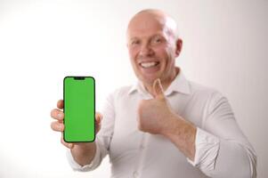 idosos branco homem 60-65 anos velho feliz e sorrir mostra fora alguma coisa em Smartphone com verde tela para Câmera. concentrando em Smartphone com chromakey Careca homem dentro uma branco camisa foto