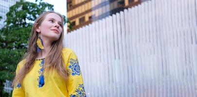 jovem lindo mulher, jovem menina dentro grande cidade dentro amarelo bordado camisa, bordado blusa, grandes Castanho cabelo, sorriso, pena brincos, amarelo e azul cores dentro roupas, ucraniano nacional traje foto