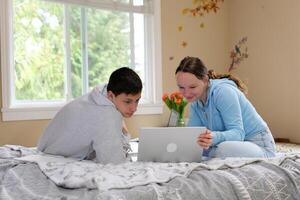 uma menina mostra interessante em formação para uma Adolescência Garoto em uma computador portátil sorridente a Garoto deram flores primeiro amor relação dentro adolescência ampla janela brilhante quarto sentar dentro quarto em cama dentro fato de treino jeans foto