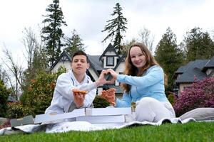 piquenique com família membros menina e Garoto mostrando coração com mãos comendo pizza dentro Parkour dentro frente do casa verão Primavera Boa Tempo com amigos e felicidade familiar e alegria ótimo ensolarado clima foto