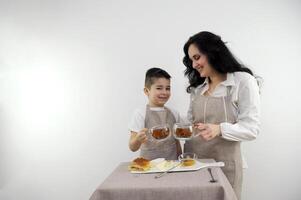 mãe e pequeno filho espalhar em queijo querida café da manhã saudável Comida em uma branco fundo beber chá cozinha avental foto