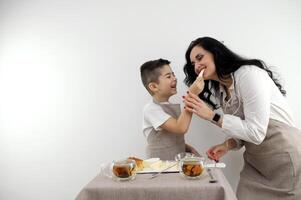 filho feeds mãe uma peça do queijo café da manhã mãe e filho experimentar queijo riso alegria agradável passatempo família sorrir cozinha avental branco fundo queijo prato foto