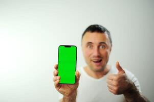 uma homem com uma verde tela em a telefone parece para dentro a quadro, Armação e mostra uma polegares acima croma chave em a telefone fechar para a Câmera publicidade espaço para texto branco fundo branco camiseta em uma homem foto