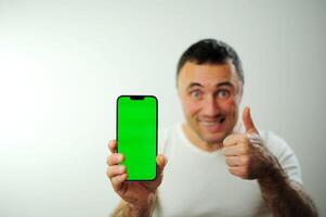 uma homem com uma verde tela em a telefone parece para dentro a quadro, Armação e mostra uma polegares acima croma chave em a telefone fechar para a Câmera publicidade espaço para texto branco fundo branco camiseta em uma homem foto