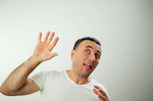 ganhar Diversão período de férias viajar diferente alegre positivo emoções do a adulto homem com cinzento Preto cabelo aberto engraçado alegre azul olhos branco camiseta cinzento fundo ruga testa bastão Fora língua foto