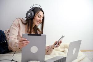 sorridente fêmea aluna desgasta sem fio fones de ouvido para estude conectados com uma skype professor, feliz jovem mulher aprende uma linguagem, ouço para uma palestra Assistir uma webinário, escrever notas, Veja às uma computador portátil foto