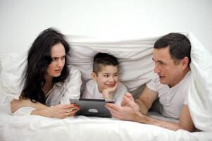 pessoas, família e tecnologia conceito mãe, pai e pequeno Garoto com tábua pc computador dentro cama às casa ou hotel sala. feliz família gastar Tempo juntos deitado debaixo branco cobertor em cama às quarto foto