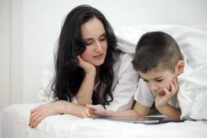 mãe e filho estão deitado debaixo a cobre às casa dentro a quarto Cuidado conectados Aprendendo Inglês curso Aprendendo tecnologia obtendo para conhecer uma tábua computador portátil Móvel apps social redes família Eu solteiro mãe foto