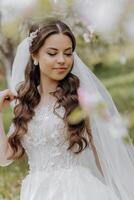 retrato do uma muito lindo noiva com grandes encaracolado cabelo e vestindo uma branco vestir dentro uma florescendo Primavera jardim foto