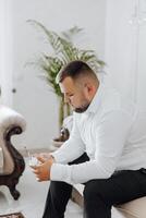 uma homem dentro uma branco camisa senta em uma sofá com uma vidro caixa para ouro Casamento argolas. a noivo é preparando para a Casamento foto