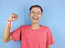 animado expressão jovem indonésio homem vestindo vermelho e branco arco de cabelo com aperto punho gesto. foto