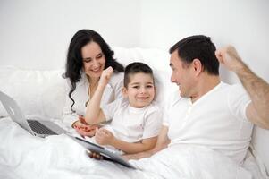 marido esposa dentro cama com pequeno filho olhando às tábua desenho animado interessante jogos As fotos rindo falando comunicação real pessoas às casa amor vida alegre pessoas espaço para texto de Anúncios social relações