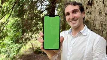 muito bonito homem francês Itália não segurando uma telefone com uma verde toque em pé dentro a madeiras dentro uma branco camisa e sorridente mostrando uma dedo em a tela pode usar para publicidade foto