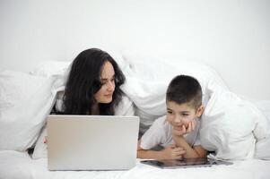 mãe e filho estão deitado debaixo a cobre às casa dentro a quarto Cuidado conectados Aprendendo Inglês curso Aprendendo tecnologia obtendo para conhecer uma tábua computador portátil Móvel apps social redes família Eu solteiro mãe foto