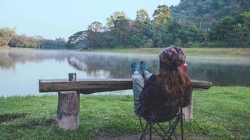 as mulheres asiáticas se sentem relaxadas levantando as pernas sobre a mesa de madeira e saboreando o café da manhã à beira do lago. viagem de acampamento foto