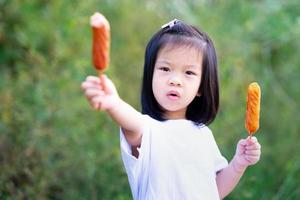 linda garota detém duas salsichas com as duas mãos. as crianças comem alimentos fáceis de comer.