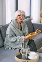 mulher sênior à moda antiga, sentada no sofá, lendo um livro em casa. educação, maduro, conceito de lazer