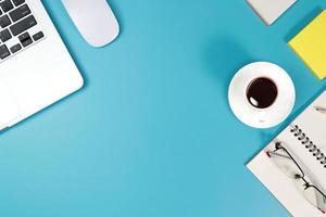 mesa plana de área de trabalho com computador laptop, material de escritório, xícara de café, telefone celular e xícara de café sobre fundo azul foto