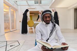 dentro a sagrado mês do Ramadã, a africano americano muçulmano homem absorta dentro lendo a piedosos Alcorão é cercado de uma pano de fundo Onde três usando hijab mulheres preparar e servir Comida para iftar foto