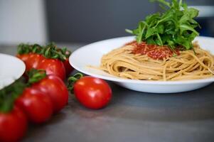 ainda vida com uma branco prato com italiano espaguete capellini com tomate molho, enfeitado com Rúcula verde folhas e ramo do vermelho maduro orgânico tomates em a cinzento cozinha mesa foto