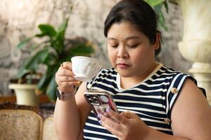 retrato do lindo plas Tamanho ásia mulher com sorridente face bebendo quente chá e usando Smartphone dentro restaurante foto