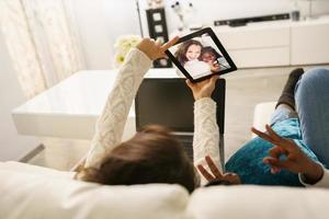 duas amigas fazendo uma selfie com um tablet digital, sentadas no sofá em casa.