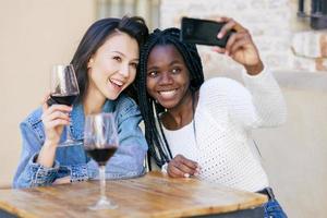 dois amigos fazendo uma selfie, sentados à mesa do lado de fora de um bar, enquanto bebem uma taça de vinho tinto.
