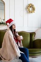 jovem sentada sozinha no sofá em uma sala de estar decorada para o natal tomando uma bebida quente