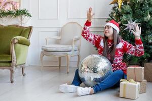 jovem com chapéu de Papai Noel segurando uma bola de discoteca no espelho dançando perto da árvore de Natal foto