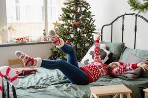 jovem de suéter vermelho e chapéu de Papai Noel deitada na cama celebrando o natal foto