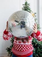 jovem com chapéu de Papai Noel segurando uma bola de espelhos em pé ao lado da árvore de Natal