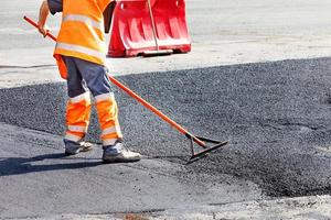 um trabalhador de serviço rodoviário alisa asfalto quente com um nível de metal manualmente ao reparar uma estrada.