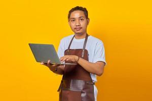 alegre e bonito jovem barista segurando um laptop e olhando para a câmera em fundo amarelo foto