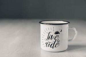 caneca esmaltada de metal branco com linha preta na borda e inscrição na mesa de madeira branca foto