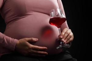 feche a mulher grávida segurando vinho tinto em um copo em fundo preto foto