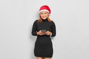 retrato de uma jovem asiática sorridente com chapéu de Papai Noel, usando telefone celular isolado sobre fundo branco foto