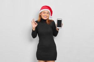 jovem asiática com chapéu de Papai Noel sorridente, segurando uma tela em branco do celular e gesticulando um sinal de ok isolado sobre o fundo branco